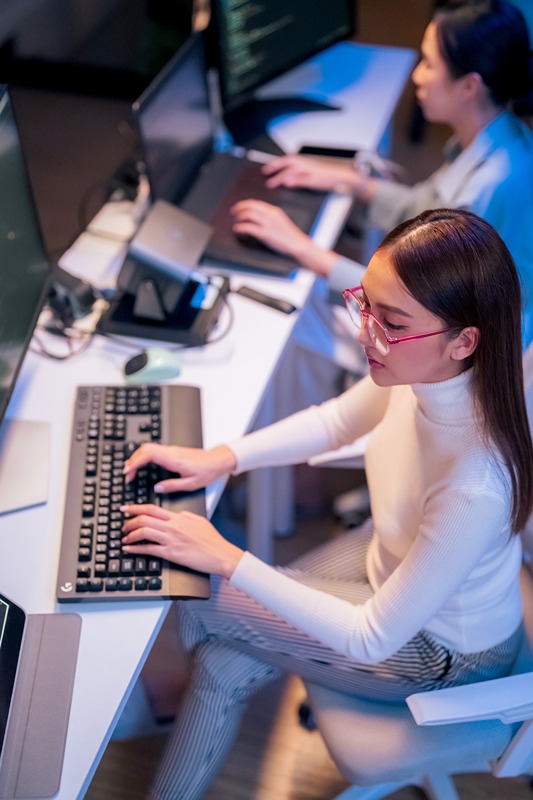 asian female programmer sits a dark tech workspace at night, fully focused on developing an online project. She writes HTML scripts and code for a modern website while contributing to automation work